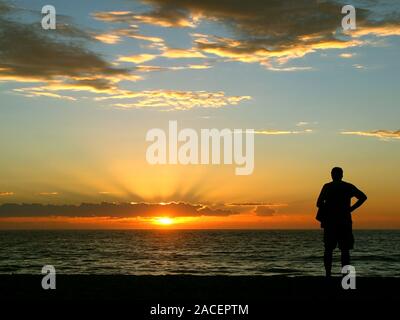 Ein älterer Mann Uhren einen wunderschönen Sonnenuntergang am Meer. Der Mann sitzt bewegungslos mit dem Rücken zur Kamera und bewundert die hellen Farben des Sonnenuntergangs. Stockfoto