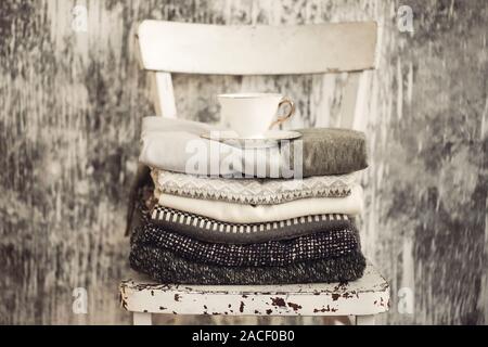 Stapel der gestrickte warme grauen und weißen Decken, Schals und Pullover aus Wolle für den Winter oder Herbst kalten Wetters auf weißen Hocker in der Nähe von weißen und grauen Wand- und Stockfoto