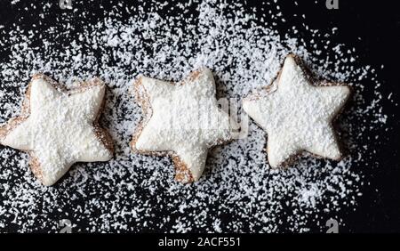 Frische Kekse in Form von Sternen mit weißem Zuckerguss und Puderzucker gegen den dunklen Hintergrund Stockfoto