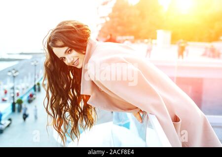 Schöne junge Mädchen mit dunklen Haaren steht in der Nähe des Meeres in einem beigen Mantel und lächelt. Sonnenuntergang auf der Sieheuntere. Stockfoto