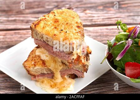 Sandwich mit Parmesan und Greyerzer Käse, Schinken, Béchamelsauce und Artisan Brot von einem Salat auf einem hölzernen Hintergrund begleitet. Der Croque Monsieur. Stockfoto
