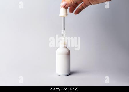 Hand der Frau mit Pipette mit Tropfen Serum oder Hyaluronsäure oder Kollagen Feuchtigkeitscreme und weiße Flasche auf grauem Hintergrund. Beauty Kosmetik consept Für feuchtigkeitsspendende Gesichtsmasken, Skin Care Solutions. Stockfoto