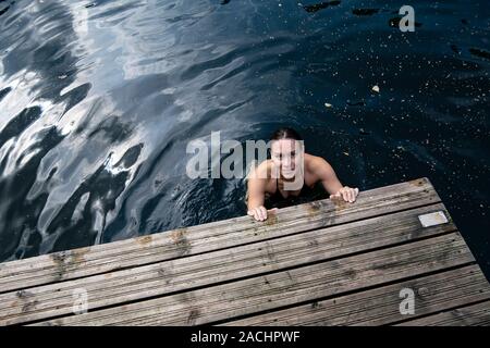 Lächelnde Frau badet in einem klaren See, sie hält ihre Hände auf den Rand eines hölzernen Pier und sieht in die Kamera. Stockfoto