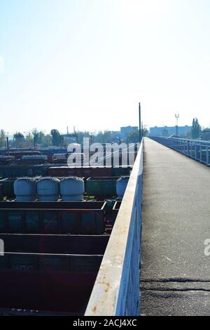 Eine schmale graue Stahlbeton Brücke über der Kreuzung von Eisenbahn- und Güterzüge durch die helle Sonne mit blauem Himmel leuchtet. Stockfoto