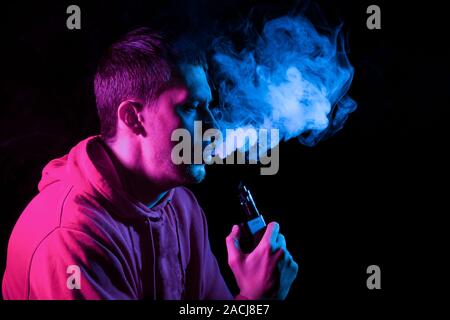 Nahaufnahme, Porträt des Gesicht von einem erwachsenen Mann atmet Blau toxischen Rauch beim Rauchen E-Zigarette und vape beleuchtet mit rosa farbiges Licht Stockfoto