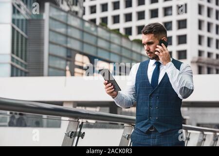 Gut aussehender Geschäftsmann spricht von Mobiltelefon und Blick auf Tablet Computer gegen Big City Stockfoto