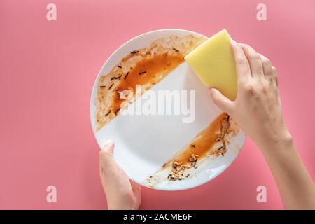 Die Frau mit dem gelben Schwamm zu reinigen und waschen Essen Flecken und Schmutz auf weißem Teller nach dem Essen Essen auf rosa Hintergrund isoliert. Reinigung Stockfoto