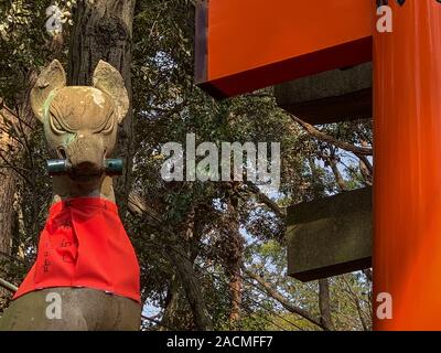 Kitsune (Fuchs) Statue trägt eine rote Latzhose und halten ein Blättern in den Mund, an den Fushimi Inari Schrein, Kyoto, Japan. Stockfoto