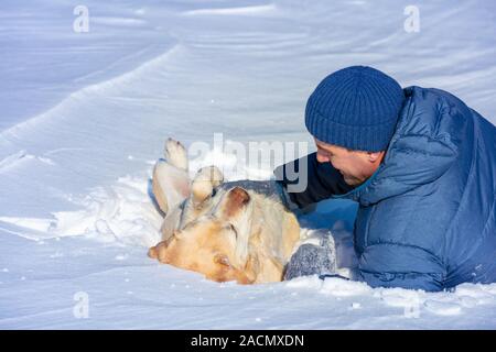 Eine glücklich lächelnde Mann mit einem Labrador Retriever Hund liegend in den Schnee im Winter. Der Hund auf dem Rücken liegend Stockfoto