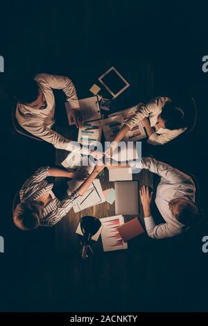 Hohe Winkel über Ansicht vertikale Foto von vier Personen Geschäftspartner sitzen Tabelle arbeiten bis spät in die Nacht Haltearme Stapel Stapel Union ein Team Seele Stockfoto