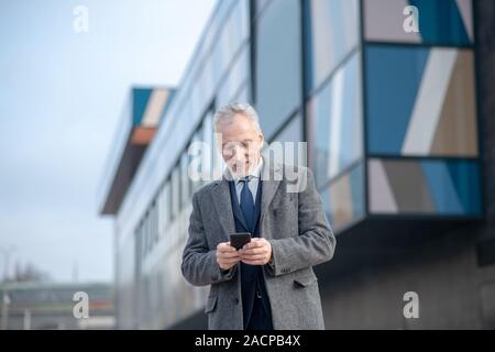 Grauhaariger Mann mit einem grauen Mantel Kontrolle sein Telefon Stockfoto