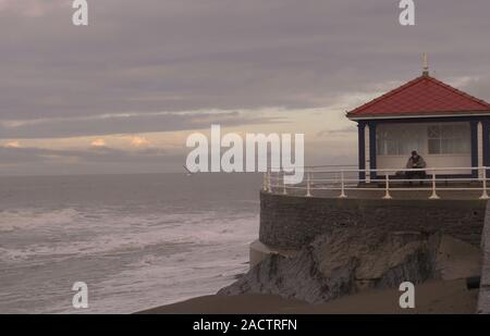 Aberystwyth Ceredigion Wales/Großbritannien, 28. November 2019: Der Mann, der ein Buch lesen am Meer öffentlichen Tierheim auf einem kalten und windigen Tag. Stockfoto