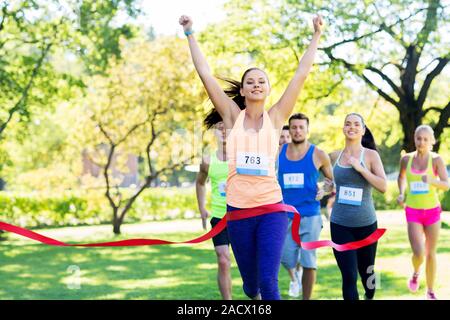 Fitness, Sport und Erfolg Konzept - glückliche Frau Rennen gewinnen und erste Kommen rotes Band über die Gruppe der Sportler läuft Marathon mit badg zu beenden Stockfoto