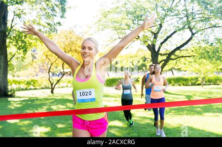 Fitness, Sport und Erfolg Konzept - glückliche Frau Rennen gewinnen und erste Kommen rotes Band über die Gruppe der Sportler läuft Marathon mit badg zu beenden Stockfoto