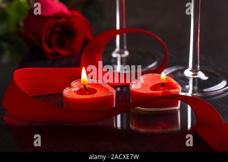 Nahaufnahme von zwei brennende Herz Valentinstag Kerzen mit roter Schleife und eine Rose im Hintergrund auf einem dunklen Tabelle Stockfoto