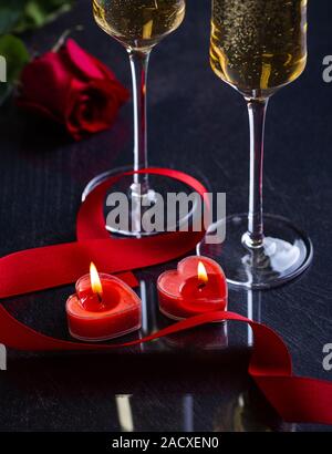 Zwei brennende Herz Valentinstag Kerzen mit roter Schleife und Gläser Champagner und Rose im Hintergrund auf einem dunklen Tabelle Stockfoto