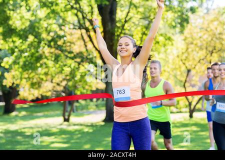 Fitness, Sport und Erfolg Konzept - glückliche Frau Rennen gewinnen und erste Kommen rotes Band über die Gruppe der Sportler läuft Marathon mit badg zu beenden Stockfoto