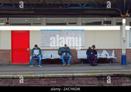 Drei junge Männer sitzen auf einem Sitz mit Blick auf ihre Smartphones beim Warten auf einen Zug bei Kingston Station Surrey, Greater London England Großbritannien Stockfoto