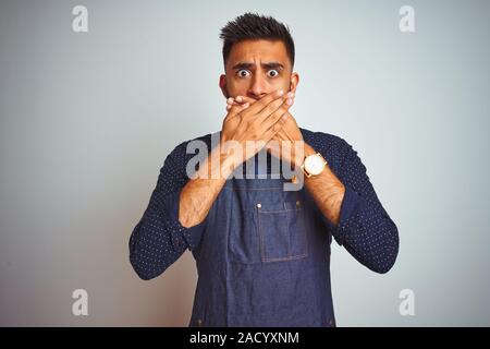 Junge indische Mitarbeiter Mann mit Schürze einheitliche stehend über isoliert weißer Hintergrund schockiert über den Mund mit Händen für Fehler. Geheime Konzept. Stockfoto