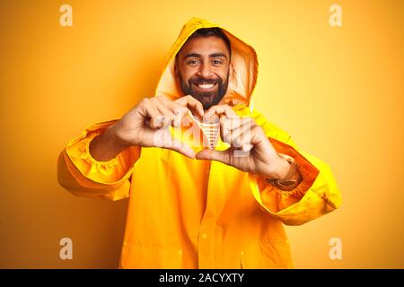 Junge inder Regenmantel mit Kapuze über isolierte gelben Hintergrund in der Liebe mit Herz und mit Hand lächelnd. Romant Stockfoto