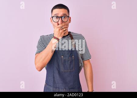 Jungen gutaussehenden Mann tragen Mitarbeiter Schürze über isolierte Hintergrund für Mund mit den Händen für den Fehler schockiert. Geheime Konzept. Stockfoto
