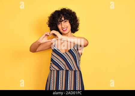 Arabische Frau mit dem lockigen Haar zu tragen gestreifte Buntes Kleid über isolierte gelben Hintergrund lächelnd in Liebe tut Herz Form mit Händen. Romantische Stunden Stockfoto