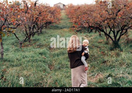 Eine Frau geht durch die herbstlichen Garten und trägt ein kleines Mädchen in die Arme. Stockfoto