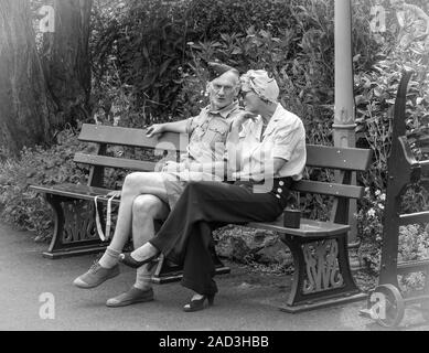 Schwarzweißfotografie, Vintage-Paar (Kostüm der 40er Jahre) auf einer Bank sitzend im Vintage-Bahnhof an der Severn Valley Heritage Railway Line. Stockfoto