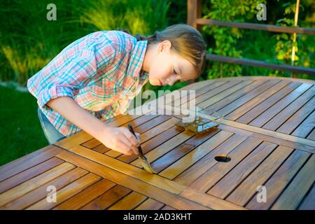 Junge hübsche Mädchen mit einem Besen Anwendung lack Farbe auf einem Holz Garten Tisch Stockfoto