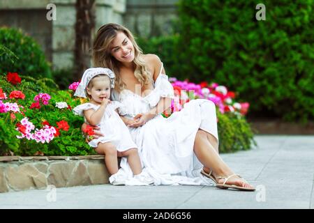 Schöne junge Mama sitzt mit ihrer Tochter in den Park. Weiße Kleider. Sie sind glücklich und Lachen. Schönes warmes Wetter. Stockfoto