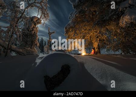 Mann neben einem Lagerfeuer in Mondbeschienene Winterlandschaft, muddus Nationalpark, Lappland, Schweden Stockfoto