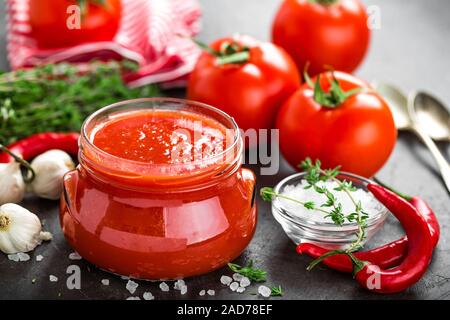 Tomatenmark, Püree in Glas und frische Tomaten auf dunklem Hintergrund. Warmes Gemüse Sauce mit Chili ant Tomaten Stockfoto