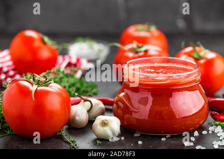 Tomatenmark, Püree in Glas und frische Tomaten auf dunklem Hintergrund. Warmes Gemüse Sauce mit Chili ant Tomaten Stockfoto