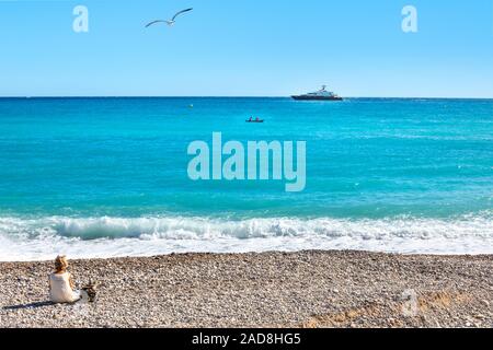Eine Frau sitzt am Strand in Menton, Frankreich an der Französischen Riviera als ein kleines Boot und größeren Boot Kreuz vor und eine Möwe fliegt über Stockfoto