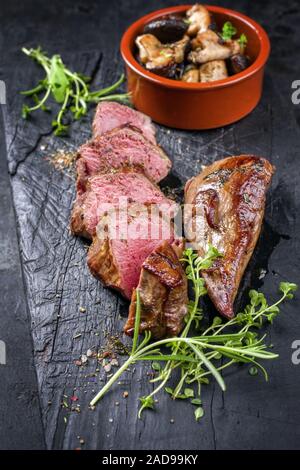 Traditionelle Grill alter geschnittenes Wildbret Rumpsteak mit Pilzen und Kräutern als Closeup auf eine verkohlte altes Board Stockfoto