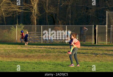 Middlefield CT USA. Apr 2019. Junge Mädchen, die kleine Schwester auf dem Rücken im Park während Jungen Baseball spielen. Stockfoto