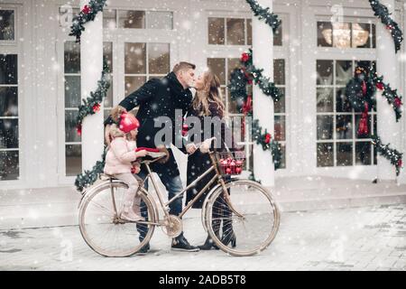 Winter Familie Gefühl der Liebe auf Vintage Fahrrad zu Weihnachten outdoor Dekoration Hintergrund posiert. Glückliche Menschen und Schwangerschaft Frau küssen während cute Dau Stockfoto