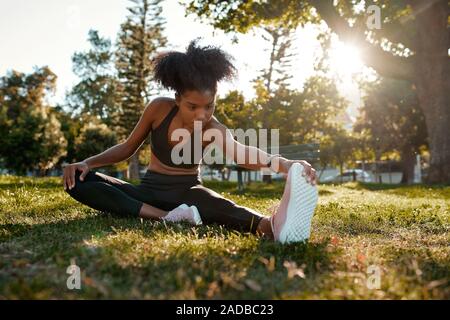 Portrait einer afrikanischen amerikanischen weiblichen Athleten ihre Beine sitzen auf der Wiese im Park - junge schwarze Frau warming up ihre Muskeln dehnen vor Stockfoto