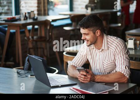 Holding smarthone und lächelnd. Erwachsene Mann sitzt im Cafe am Tag und mit dem Laptop für die Fernbedienung Stockfoto