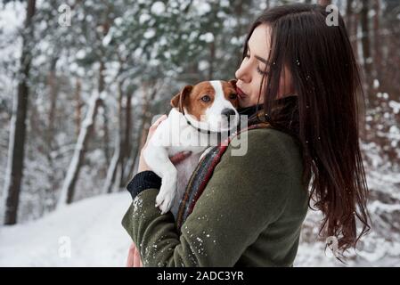 Das Küssen das Haustier, während Er ihn auf den Händen. Smiling brunette Spaß beim Spaziergang mit ihrem Hund im Winter Park Stockfoto