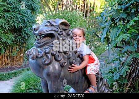 Glücklich lächelnde Baby Mädchen reiten eine Statue des mythologischen Löwe im Königlichen Botanischen Garten in Sydney, Australien Stockfoto