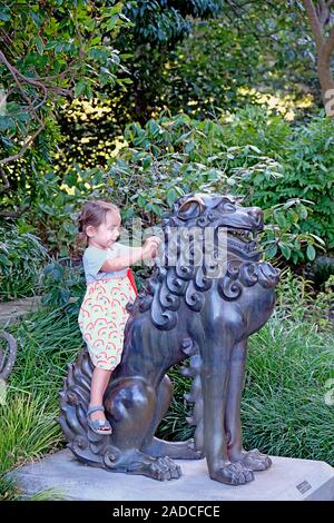 Glücklich lächelnde Baby Mädchen reiten eine Statue des mythologischen Löwe im Königlichen Botanischen Garten in Sydney, Australien Stockfoto