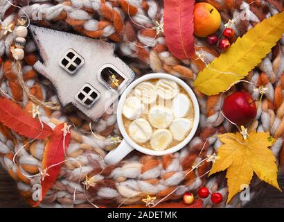 Herbst Komposition. Tasse Kakao mit Marshmallows, Holzhütte, bunte Blätter und Schal auf Tisch. Stockfoto