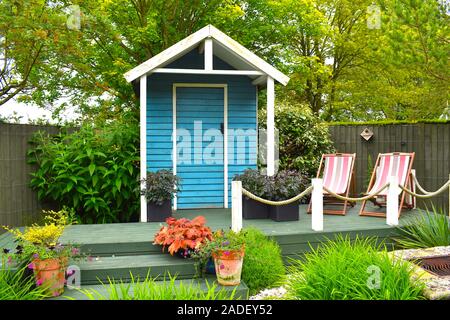 Holzblau lackierte Gartenwerkzeuge Lagerhalle zwischen grünem Gras, Topfpflanzen und rosafarbenen Deckstühlen im Sommer in der englischen Landschaft. GROSSBRITANNIEN Stockfoto