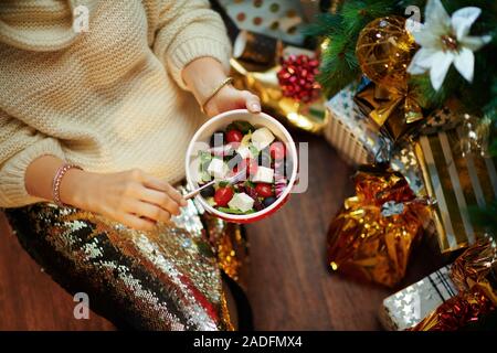 Nahaufnahme auf mittleren Alter Hausfrau in Gold Pailletten Rock und weißen Pullover unter dem geschmückten Weihnachtsbaum in der Nähe des heutigen Boxen gesund essen Salat. Stockfoto