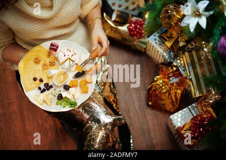 Nahaufnahme auf junge hausfrau in Gold Pailletten Rock und weißen Pullover unter dem geschmückten Weihnachtsbaum in der Nähe des heutigen Boxen essen Käse und Plattenteller. Stockfoto