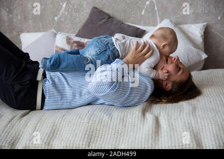 Glückliche Junge kaukasier Familie im Studio. Mutter halten und umarmte seine Tochter in den Händen. Liegende Frau auf dem Bett. Frau lächelnd. Kleinkind liegen auf M Stockfoto