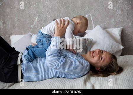 Glückliche Junge kaukasier Familie im Studio. Mutter halten und umarmte seine Tochter in den Händen. Liegende Frau auf dem Bett. Frau lächelnd. Kleinkind liegen auf M Stockfoto