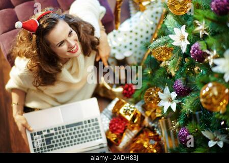 Obere Ansicht von Happy trendy Hausfrau mit langen brünetten Haar in Gold Pailletten Rock und weißen Pullover unter dem geschmückten Weihnachtsbaum in der Nähe des heutigen Boxen Stockfoto