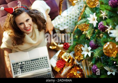 Obere Ansicht von Happy trendy mittleren Alter Frau mit langen brünetten Haar in Gold Pailletten Rock und weißen Pullover unter dem geschmückten Weihnachtsbaum in der Nähe des heutigen Stockfoto